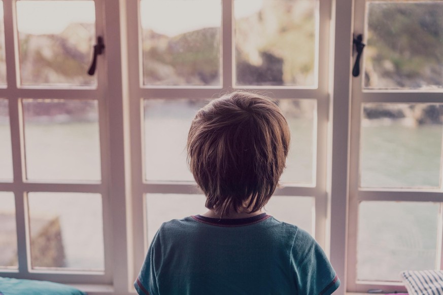 A young boy looking out of the window of a cottage by the coast.