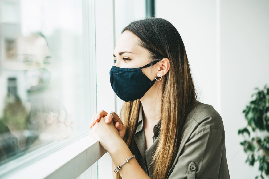 A sad girl in a face mask looks out the window from home.