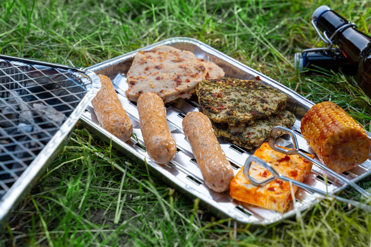 Kammlach, Bavaria, Germany - June 25, 2025: Vegetarian barbecue in the garden: disposable grill with charcoal, barbecue food made from plant-based meat substitutes and cold beer on the summer meadow r ...